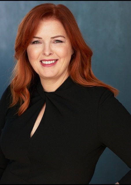 Smiling woman with red hair wearing a black top against a blue background.